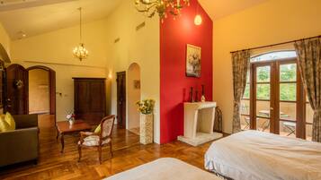 A hotel room with a wooden floor, a red accent wall, a chandelier, a fireplace, and a view through large windows.