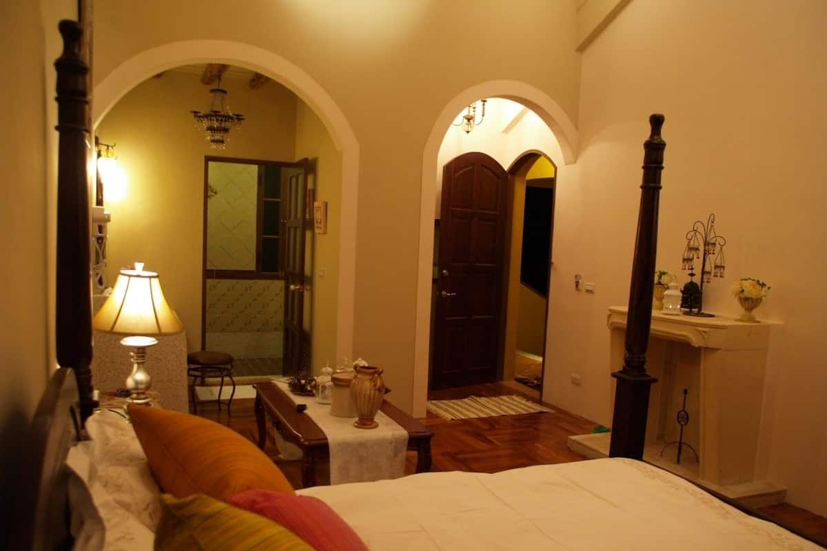 A bedroom with a large bed, a nightstand with a lamp, a chair, and a small table with decorative items. There are two doors leading to other rooms.