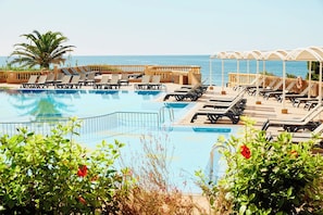 A pool area with lounge chairs and umbrellas, surrounded by greenery and a view of the sea.