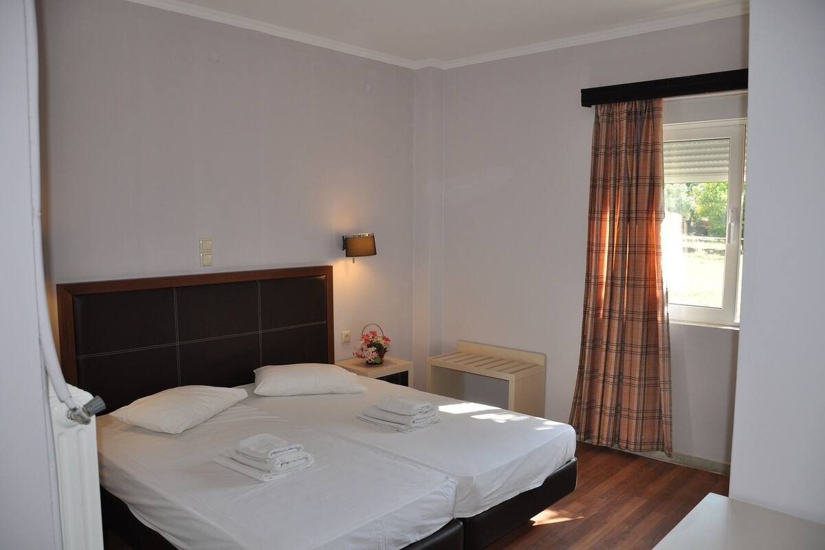 A neatly made bed with white linens and towels, a bedside table with a lamp, and a window with plaid curtains.