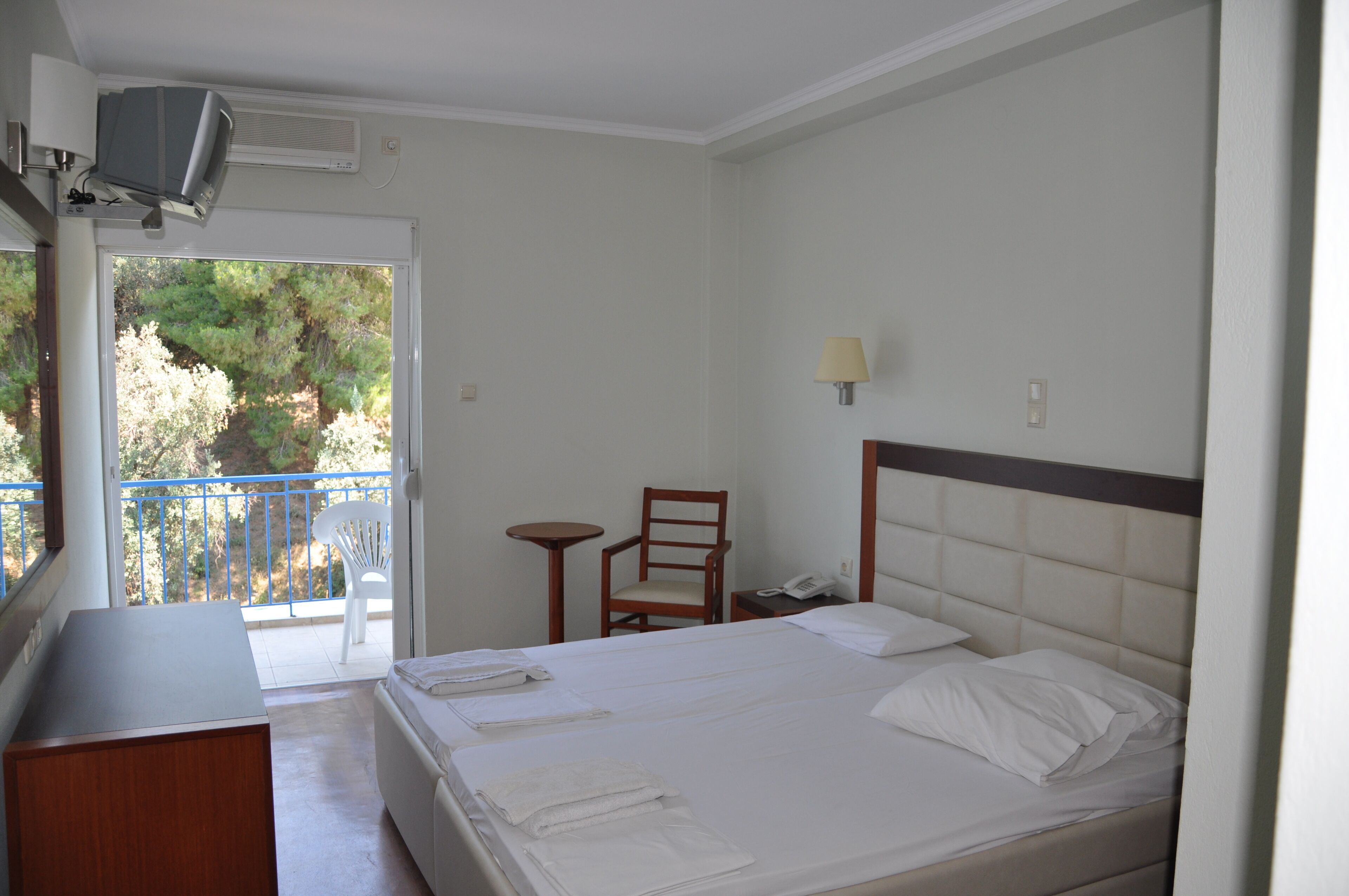 A hotel room with a bed, a desk, a chair, a small table, and a balcony with a view of trees.
