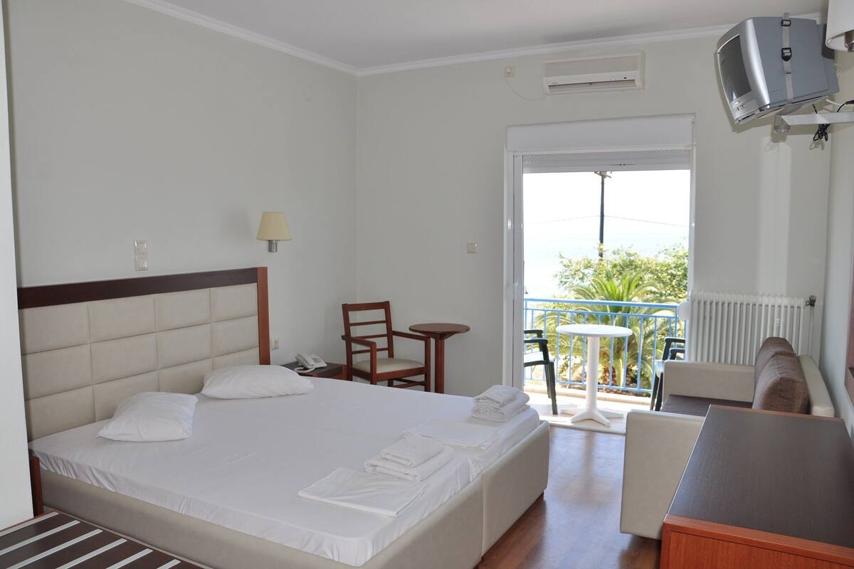 A hotel room with a bed, a desk, a chair, a small table, and a balcony with a view.