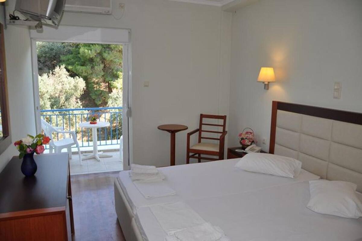 A hotel room with a bed, a small table with a vase of flowers, a chair, and a balcony with a view of trees.