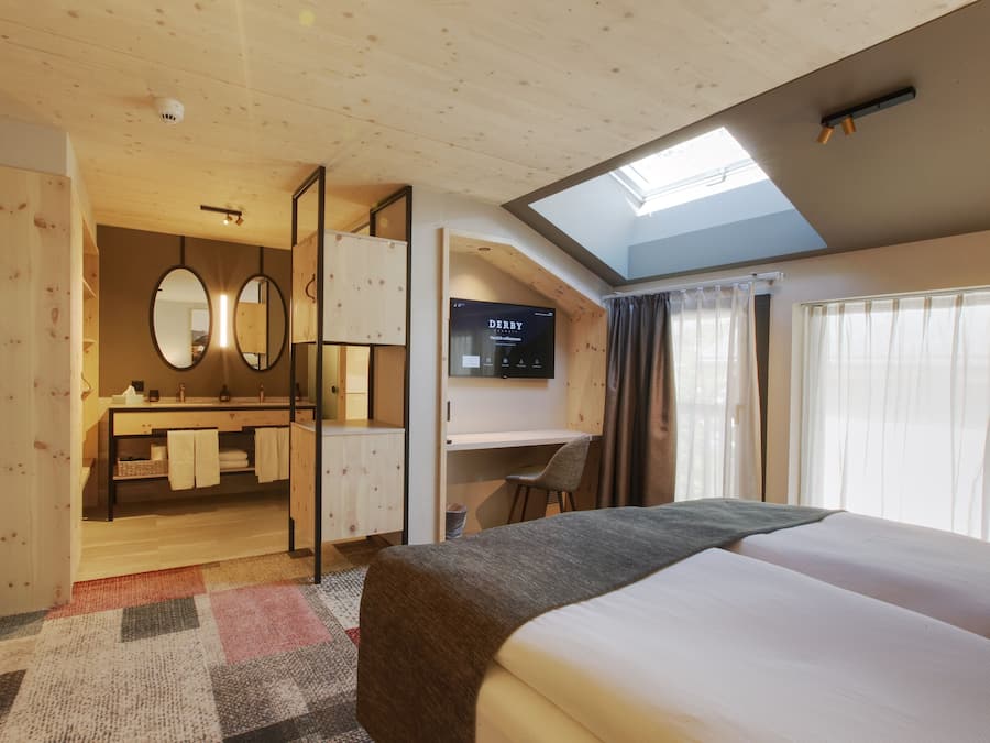 Habitación de hotel con una cama grande, un escritorio, un espejo y un televisor mostrando 'DERBY'.