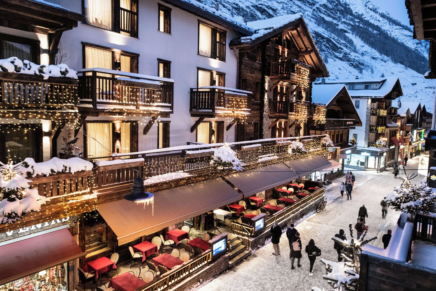 A snow-covered outdoor dining area with multiple tables and chairs, a wooden building with balconies, and a mountainous backdrop.
