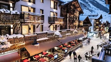 A snow-covered outdoor dining area with multiple tables and chairs, a wooden building with balconies, and a mountainous backdrop.