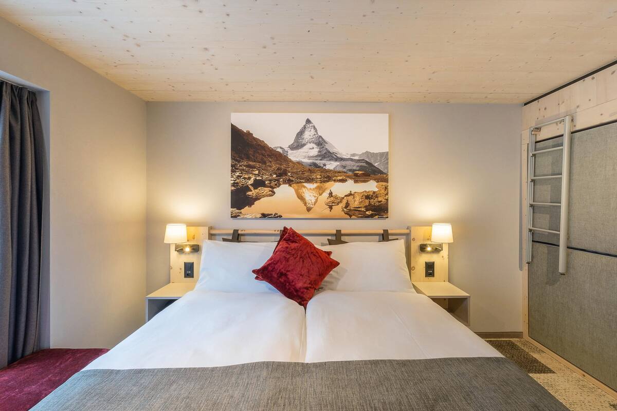 Habitación de hotel con cama, dos lámparas de noche, almohada roja, cuadro de una montaña y una escalera apoyada en la pared.