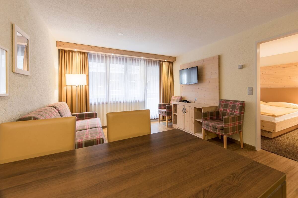 Habitación de hotel con una mesa de comedor de madera, dos sillas, un sofá, un escritorio con una silla, un televisor montado en la pared y una cama.