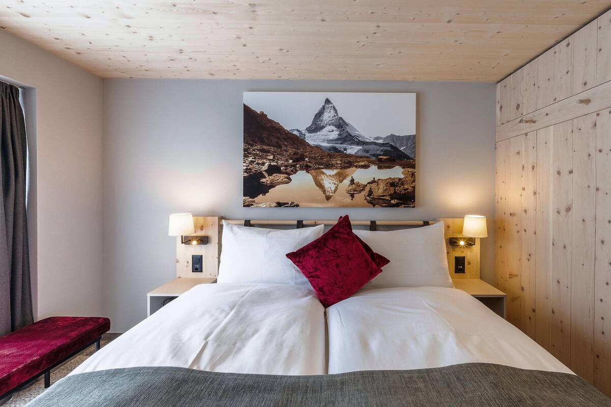 Una habitación de hotel con una cama, dos lámparas de noche, un almohadón rojo, un cuadro de una montaña y paneles de madera en las paredes.
