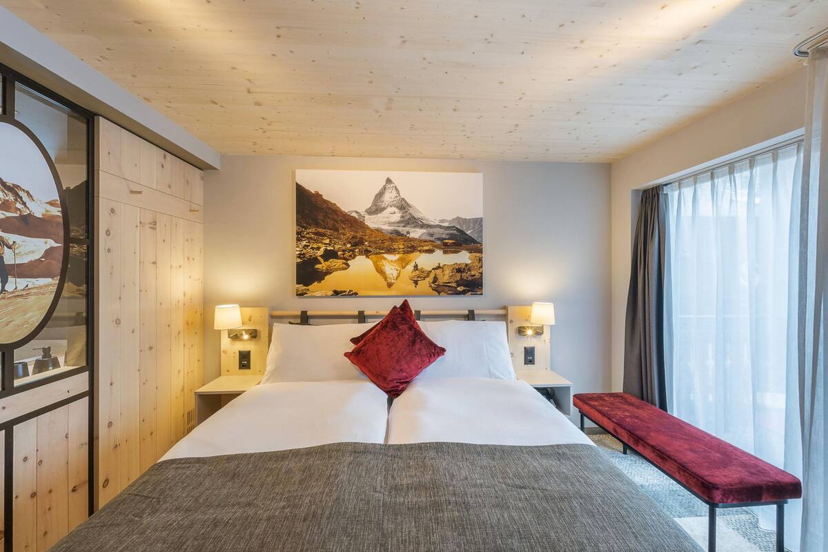 Habitación de hotel con una cama grande, dos lámparas de noche, una almohada roja, un banco y un cuadro de un paisaje montañoso en la pared.
