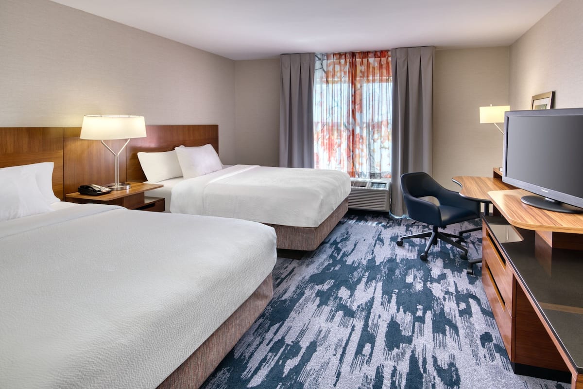 Quarto de hotel com duas camas, uma escrivaninha, uma televisão e uma janela com cortinas.