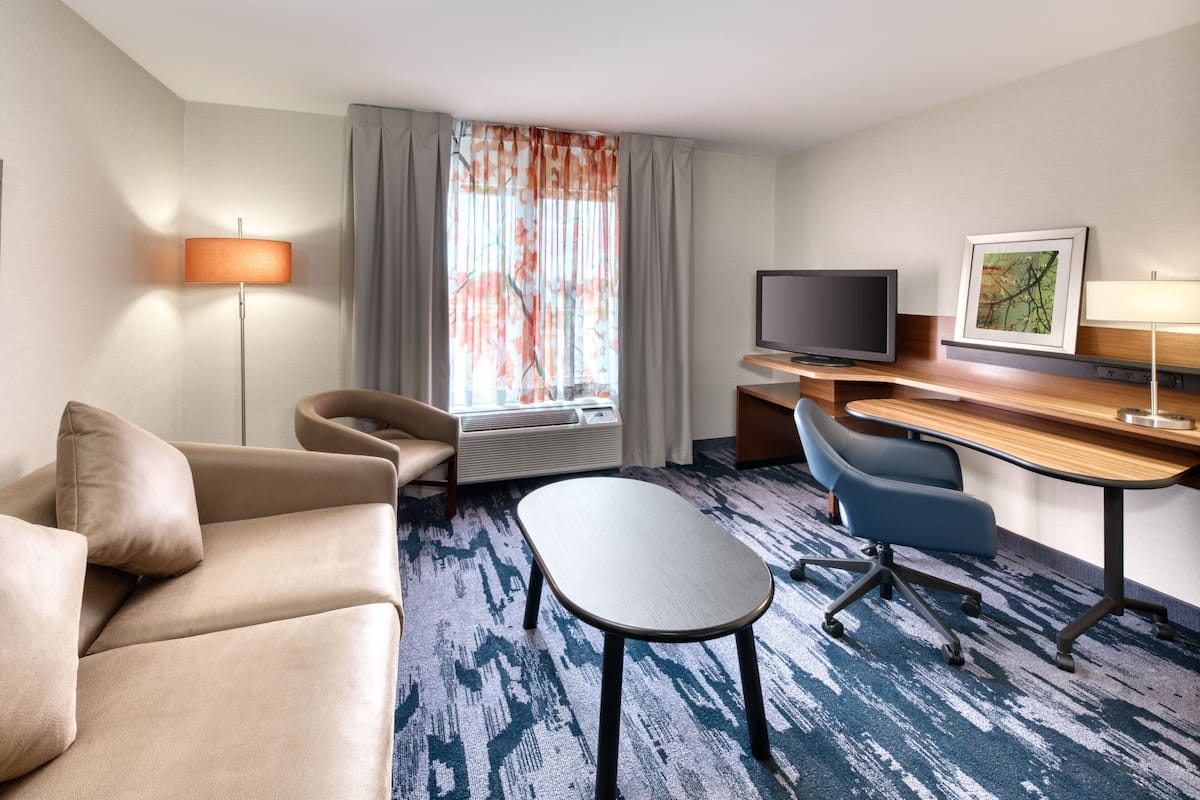 Quarto de hotel com sofá, escrivaninha, cadeira, televisão e janela com cortinas.