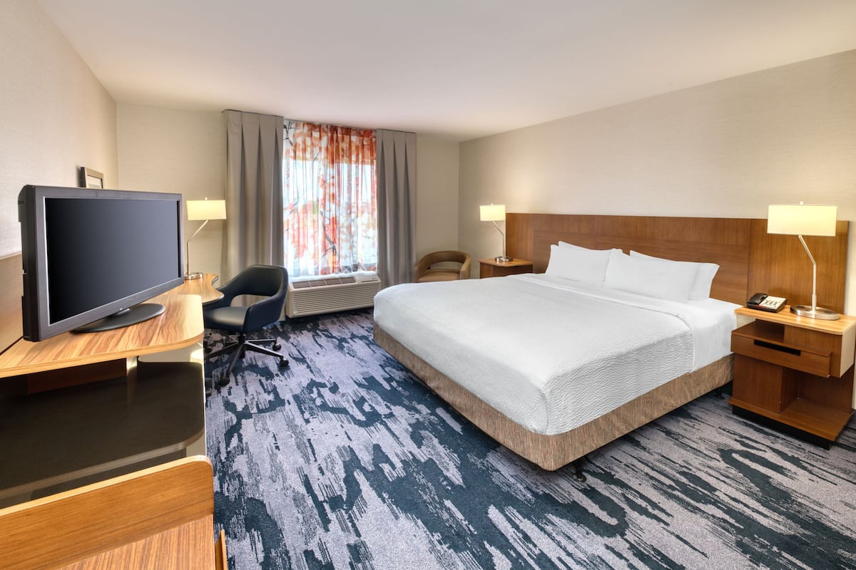Quarto de hotel com uma cama grande, uma mesa de trabalho com uma televisão de tela plana, uma cadeira, um abajur e uma janela com cortinas.