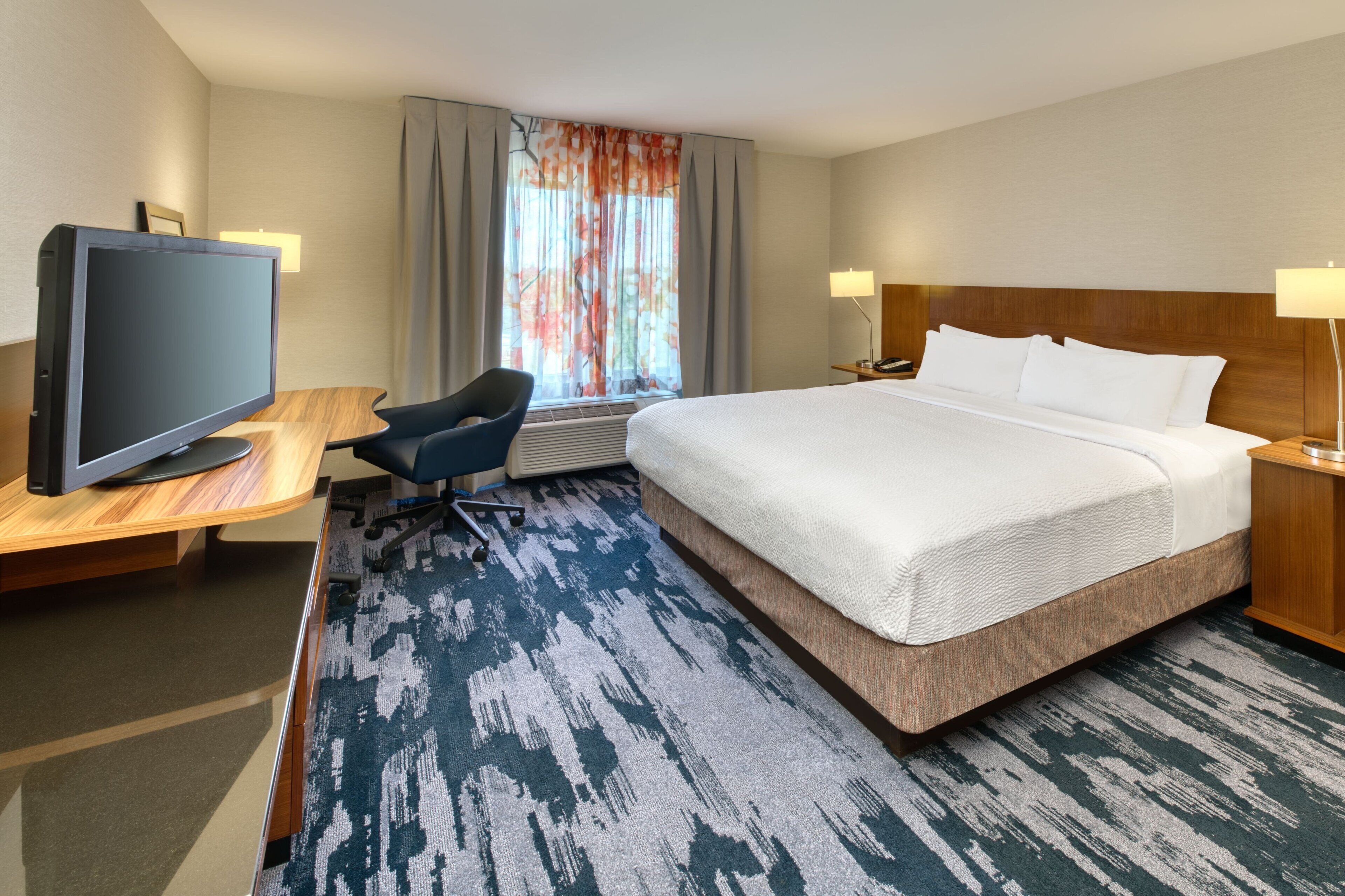 Habitación de hotel con una cama grande, un escritorio con televisor, una silla y un ventanal con cortinas.