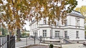 A large, white building with a symmetrical facade, a central entrance with steps, and a small garden area with gravel and a few plants. - Hotel Red Baron (Swidnica)