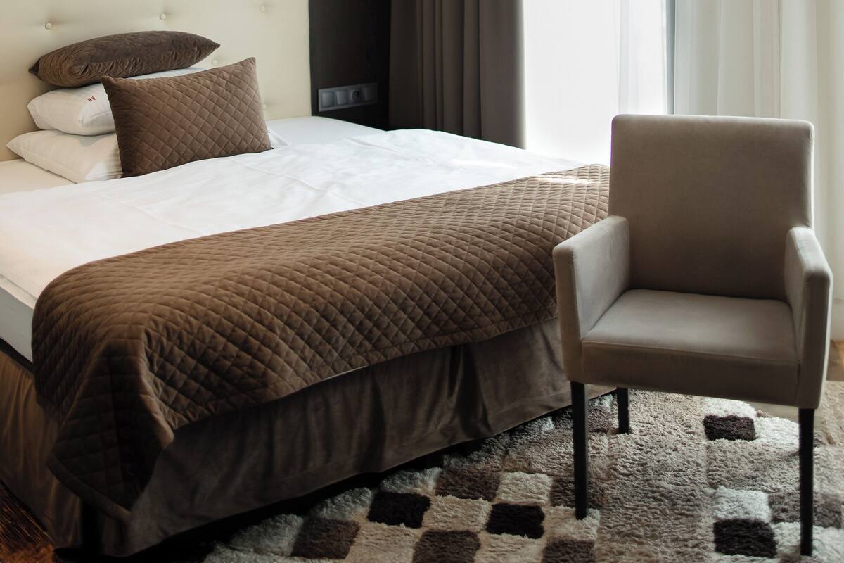 A hotel room with a bed, a chair, a nightstand, a lamp, and a window with curtains.