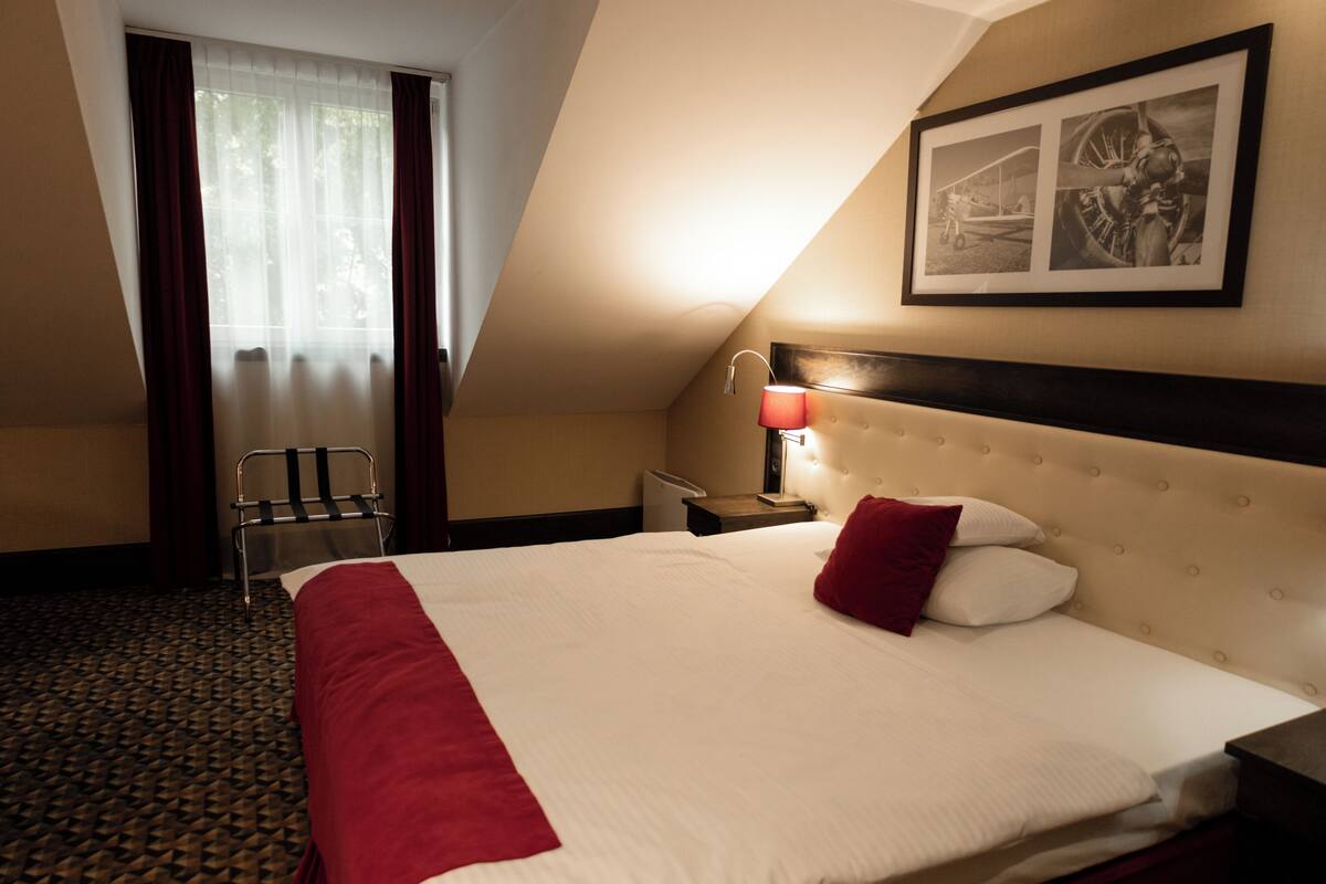 A hotel room with a large bed, a nightstand with a lamp, a chair, and a window with curtains.