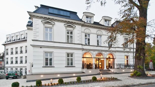 A white building with a classic architectural style, featuring a central entrance with arched windows and a sign that reads "Red Barn Inn." - Hotel Red Baron (Swidnica)