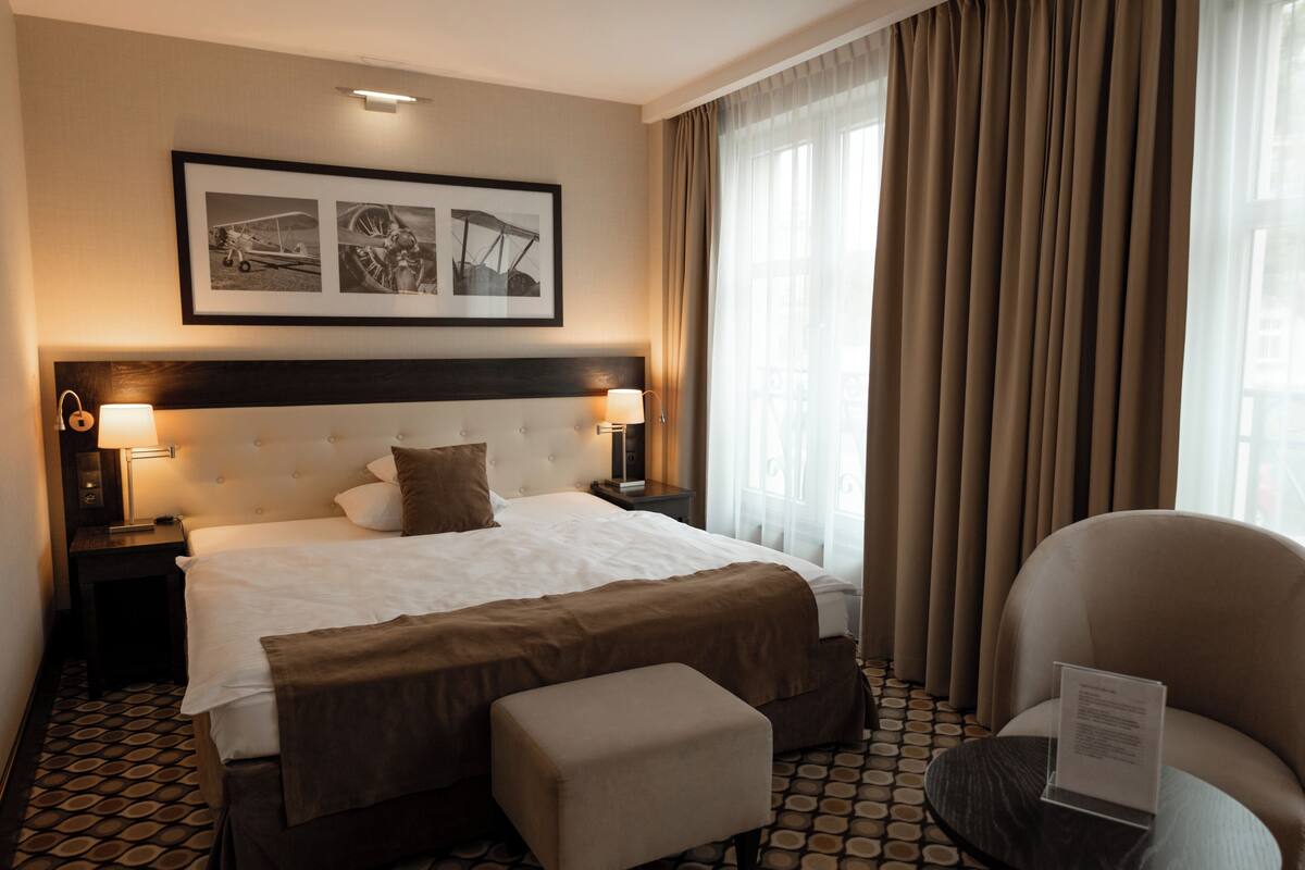 A hotel room with a large bed, two bedside lamps, a headboard, a framed picture above the bed, a window with curtains, and a chair with a sign on it.