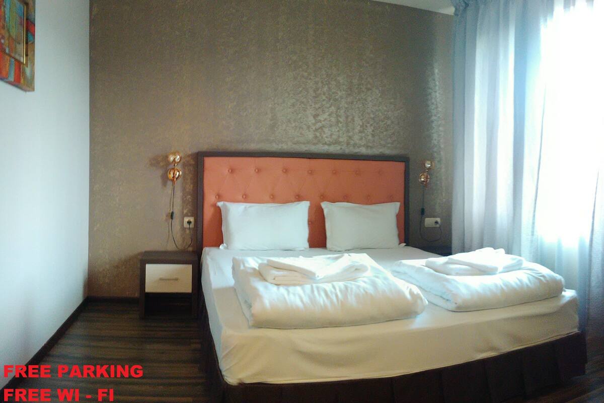 A hotel room with a neatly made bed, a nightstand, and a wall-mounted lamp.