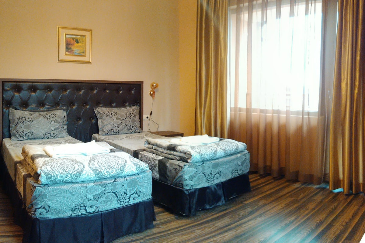 A hotel room with a large bed, two smaller beds, a headboard, a nightstand, a lamp, a painting on the wall, and a window with curtains.