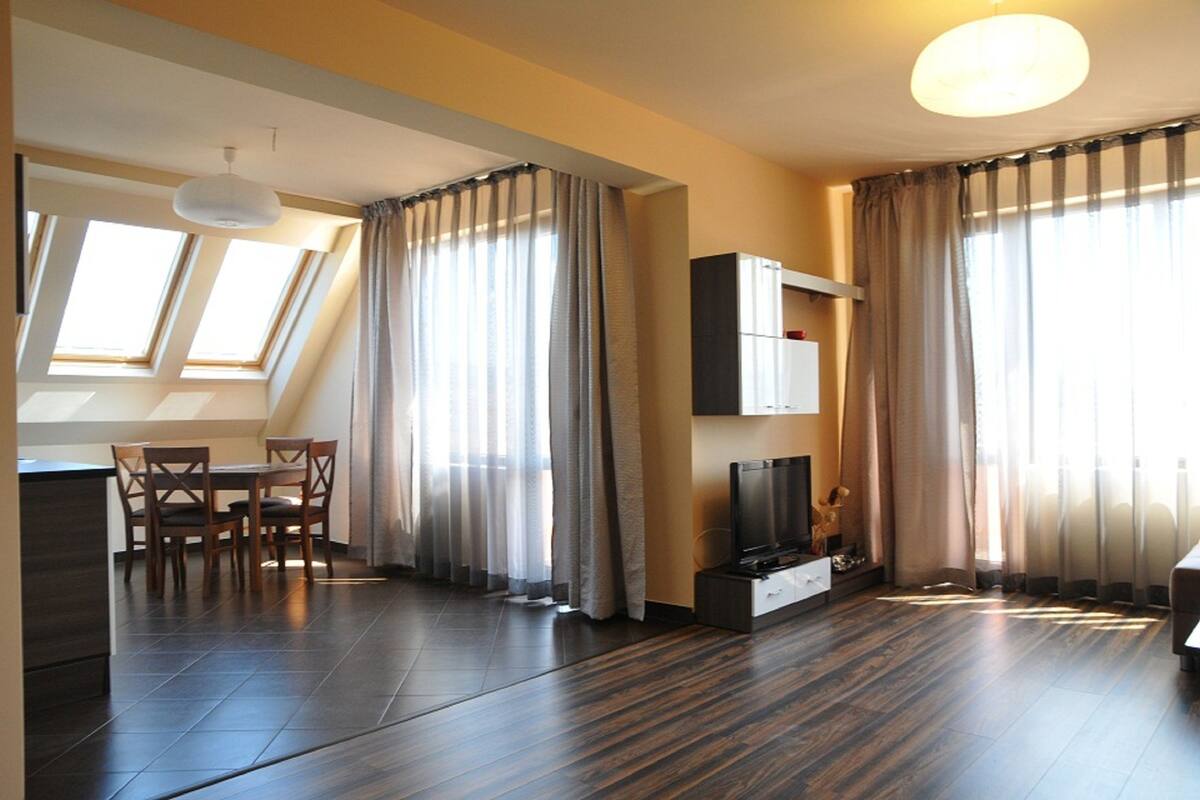 A modern living room with a dining area, large windows with curtains, a flat-screen TV, and a ceiling light.