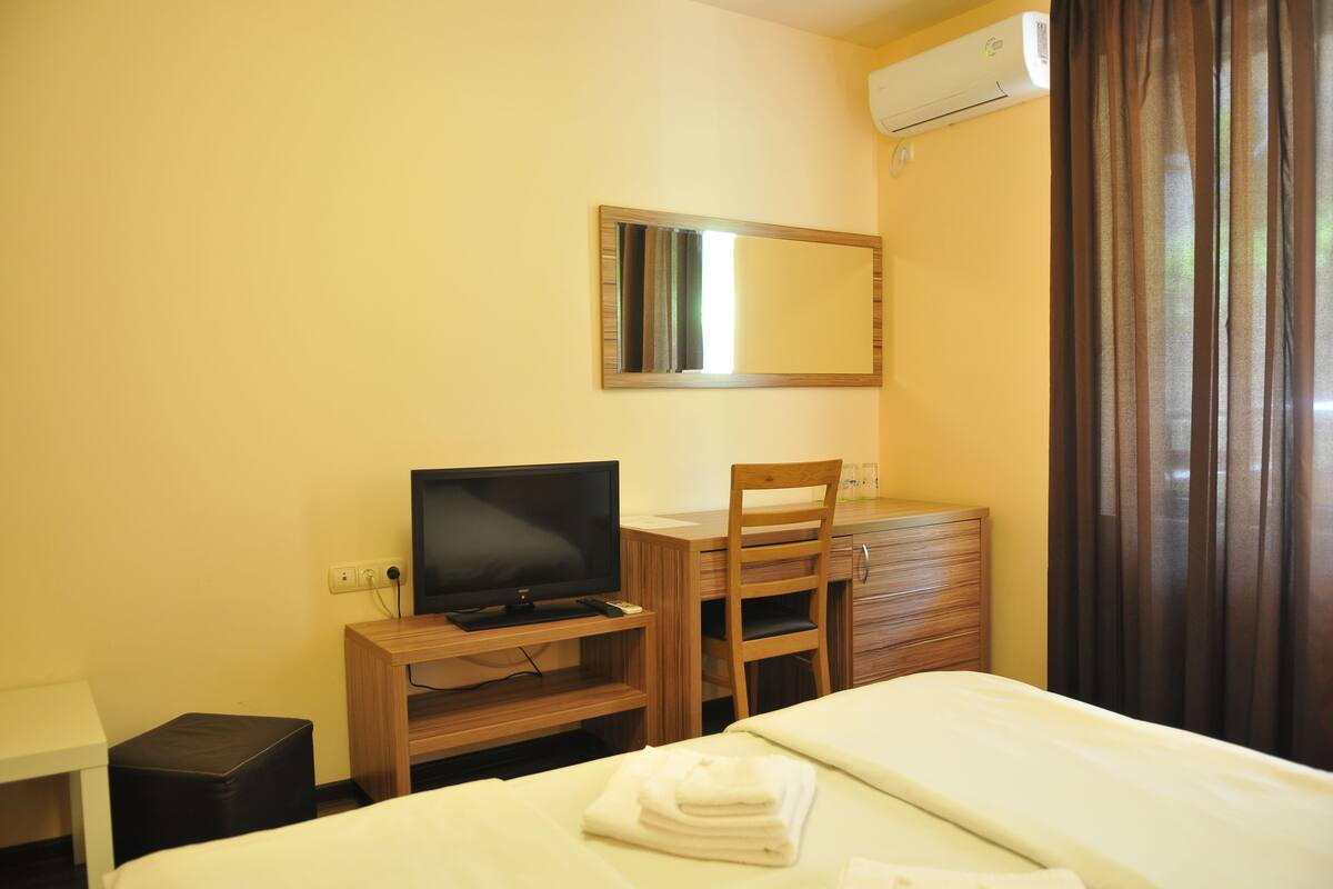 A hotel room with a bed, a desk with a television, a chair, a dresser, and an air conditioning unit.