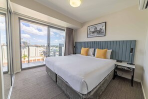 A hotel room with a large bed, a nightstand, a desk, and a view of the city through a sliding glass door. - Quest Rockhampton (Rockhampton)