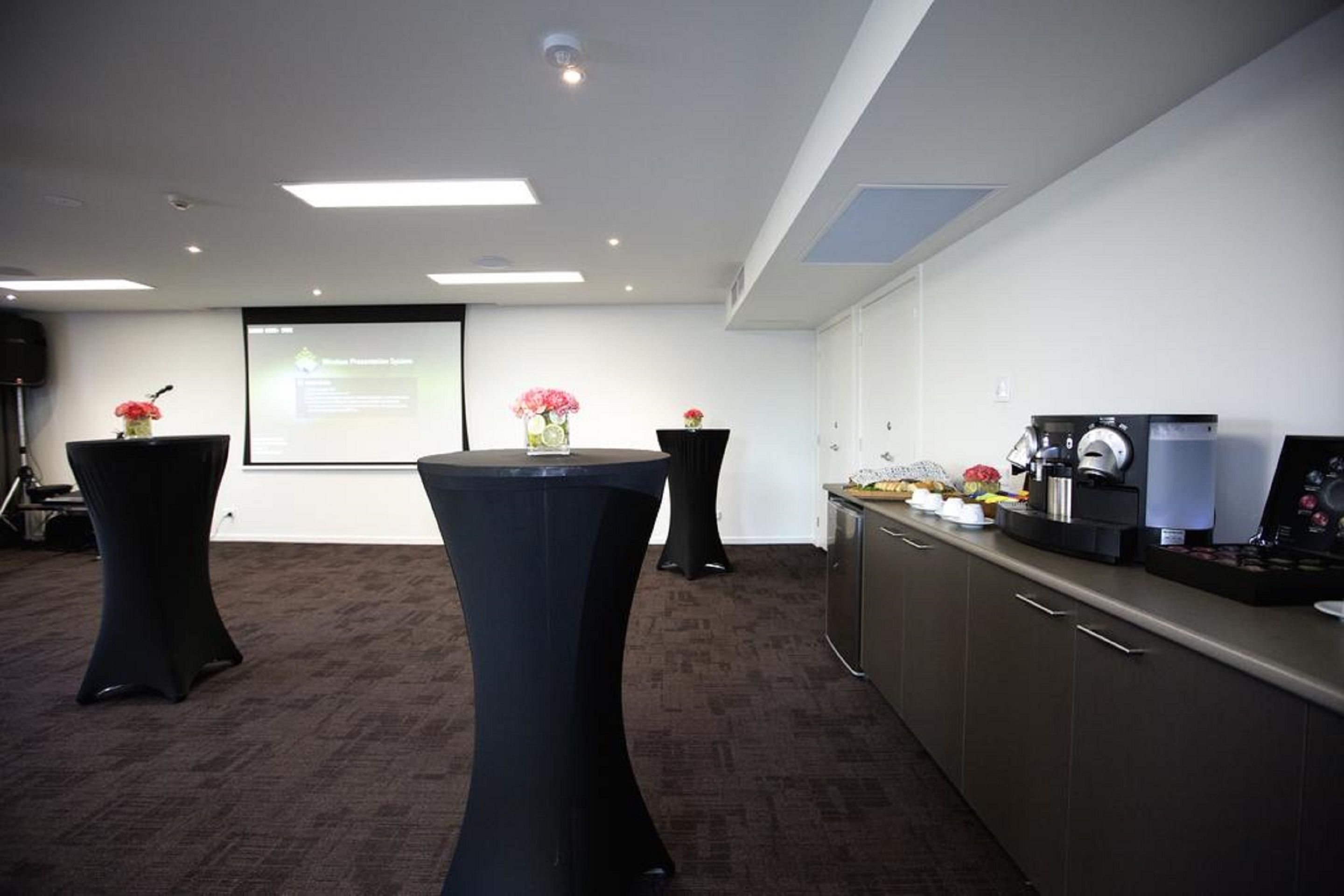 A conference room with a projector screen, two black tables with floral centerpieces, a coffee machine, and a kitchenette area.