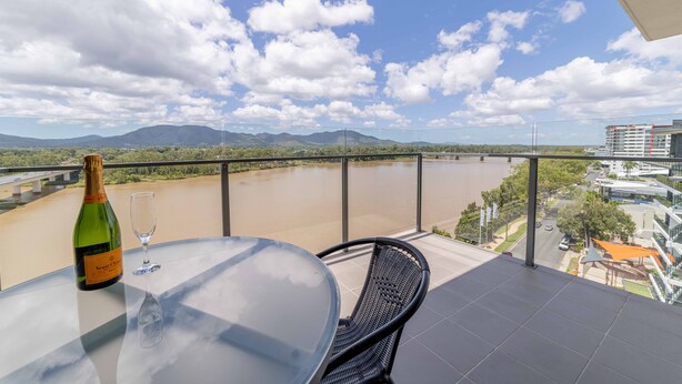 A balcony with a table, a bottle of champagne, and a glass. The view includes a river, mountains, and buildings.