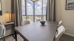 A modern hotel room with a large window offering a city view, a dining table with chairs, a desk with a lamp, and a framed picture on the wall.