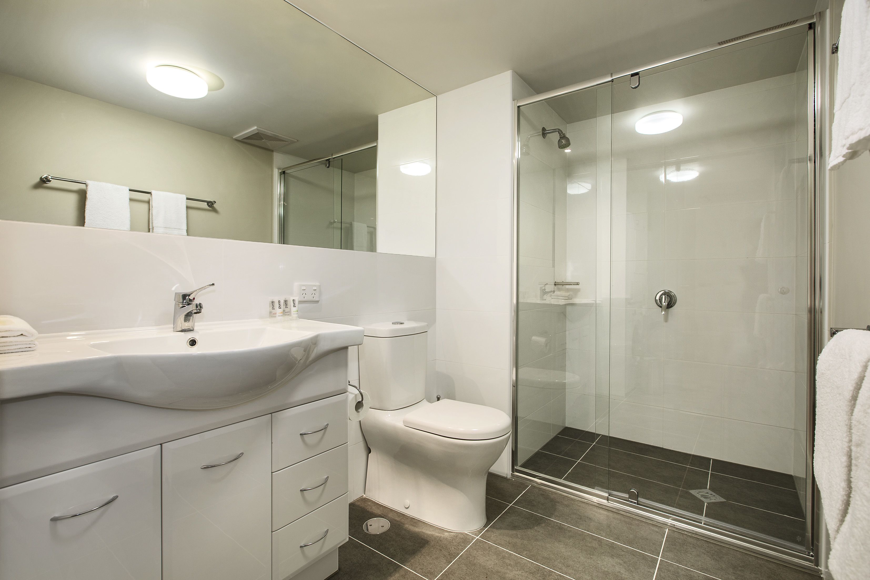 Une salle de bain moderne avec un lavabo, un meuble-lavabo et des toilettes blancs, ainsi qu’une douche séparée par une paroi de verre.