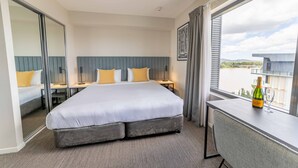 A hotel room with a large bed, a desk, a chair, a bottle of champagne, and a view of the city.