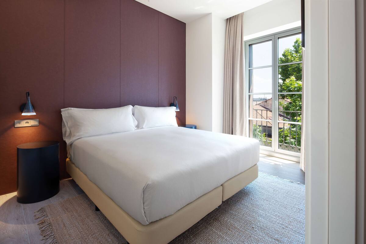 Een moderne hotelkamer met een groot bed, twee bedlampjes, uitzicht op de omgeving door een groot raam en een donkere wand.