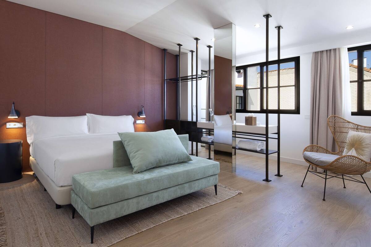 Een moderne slaapkamer met een groot bed, een groene poef, een rieten stoel en een badkamer zichtbaar door een glazen wand.