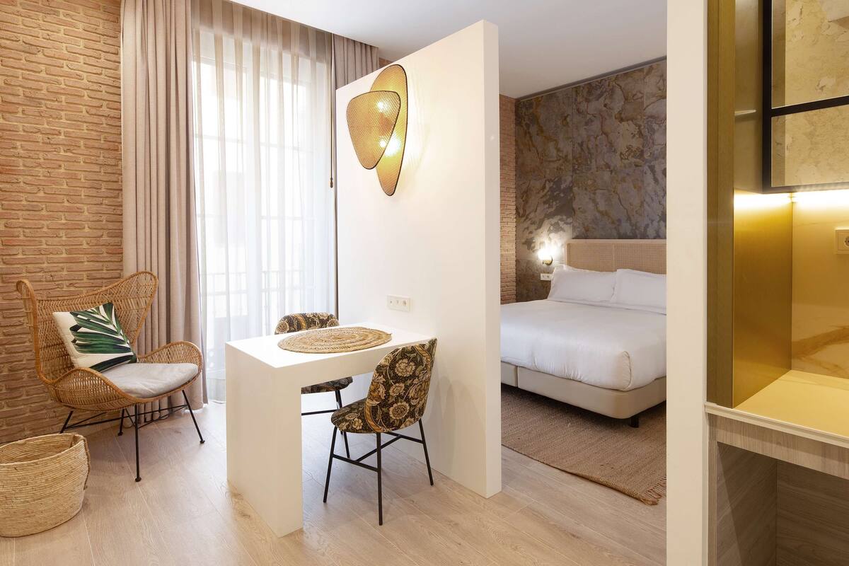 Een moderne hotelkamer met een bakstenen muur, een zithoek met rieten meubels, een bed met hoofdeinde en een wandlamp.