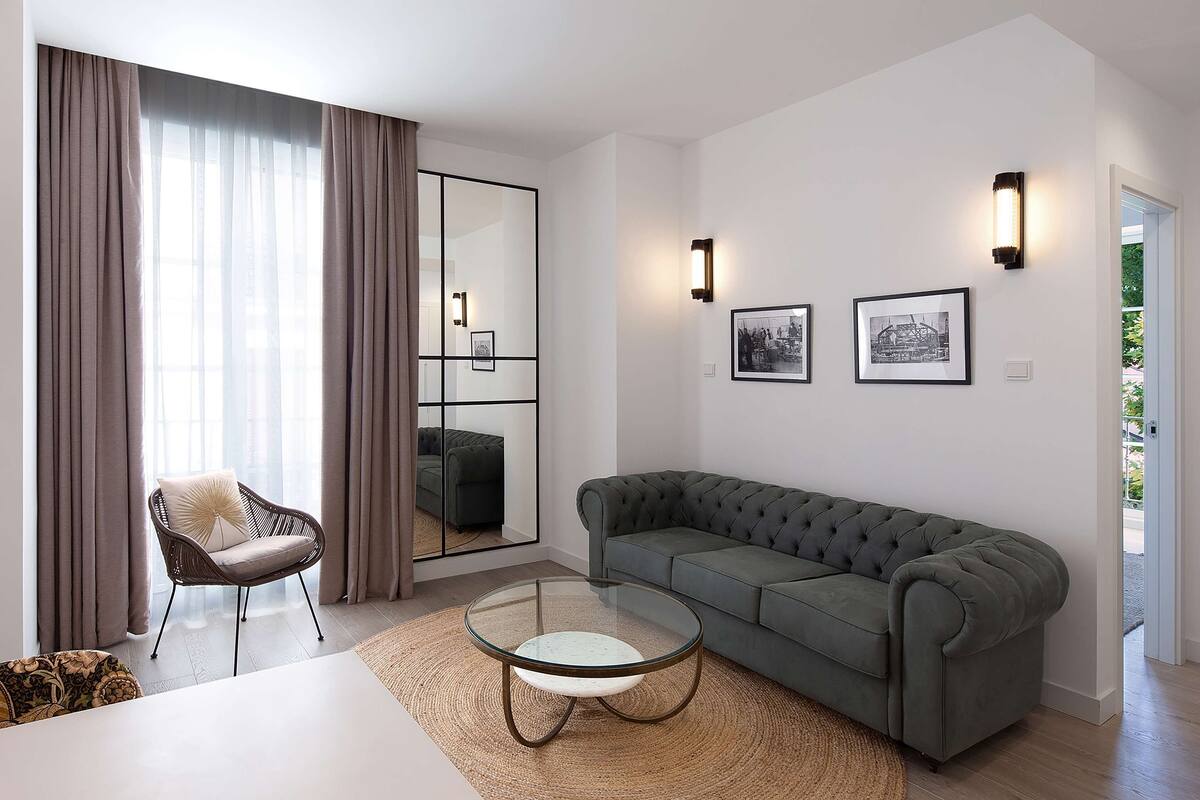 Een moderne woonkamer met een modulaire bank, een glazen salontafel, een rieten fauteuil en een groot raam met gordijnen.