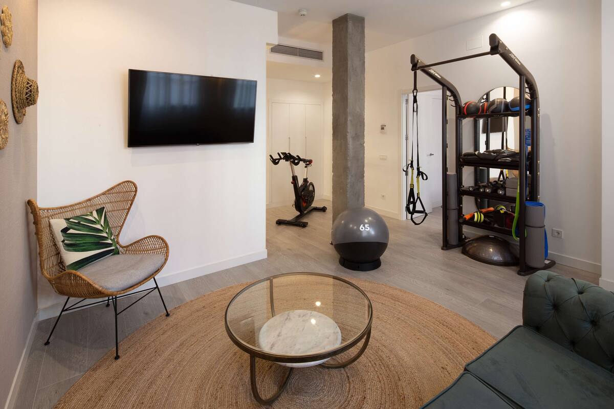Een moderne woonkamer met een glazen salontafel, een rieten fauteuil, een groene bank, een aan de muur bevestigde televisie en een thuisgym.