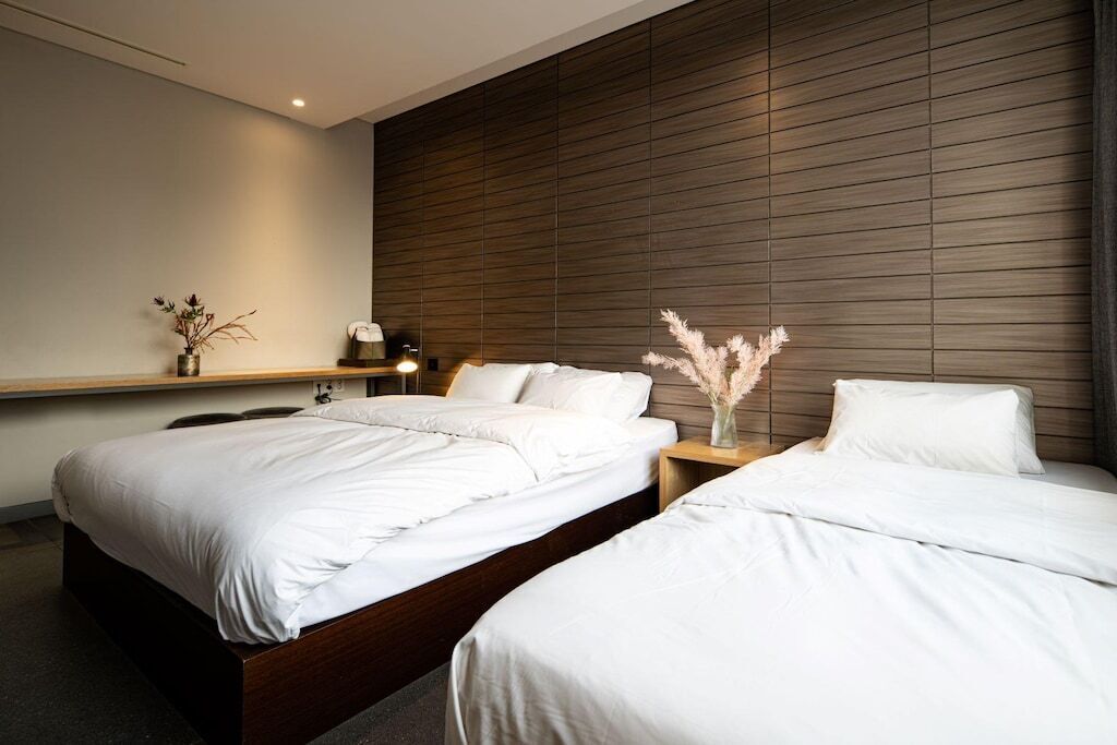 Hotelkamer met twee bedden, een houten hoofdbord, een nachtkastje met een lamp en een vaas met gedroogde bloemen.