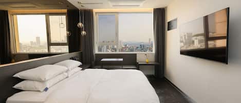 A modern hotel room with a large bed, a desk, a chair, and a large window offering a city view.