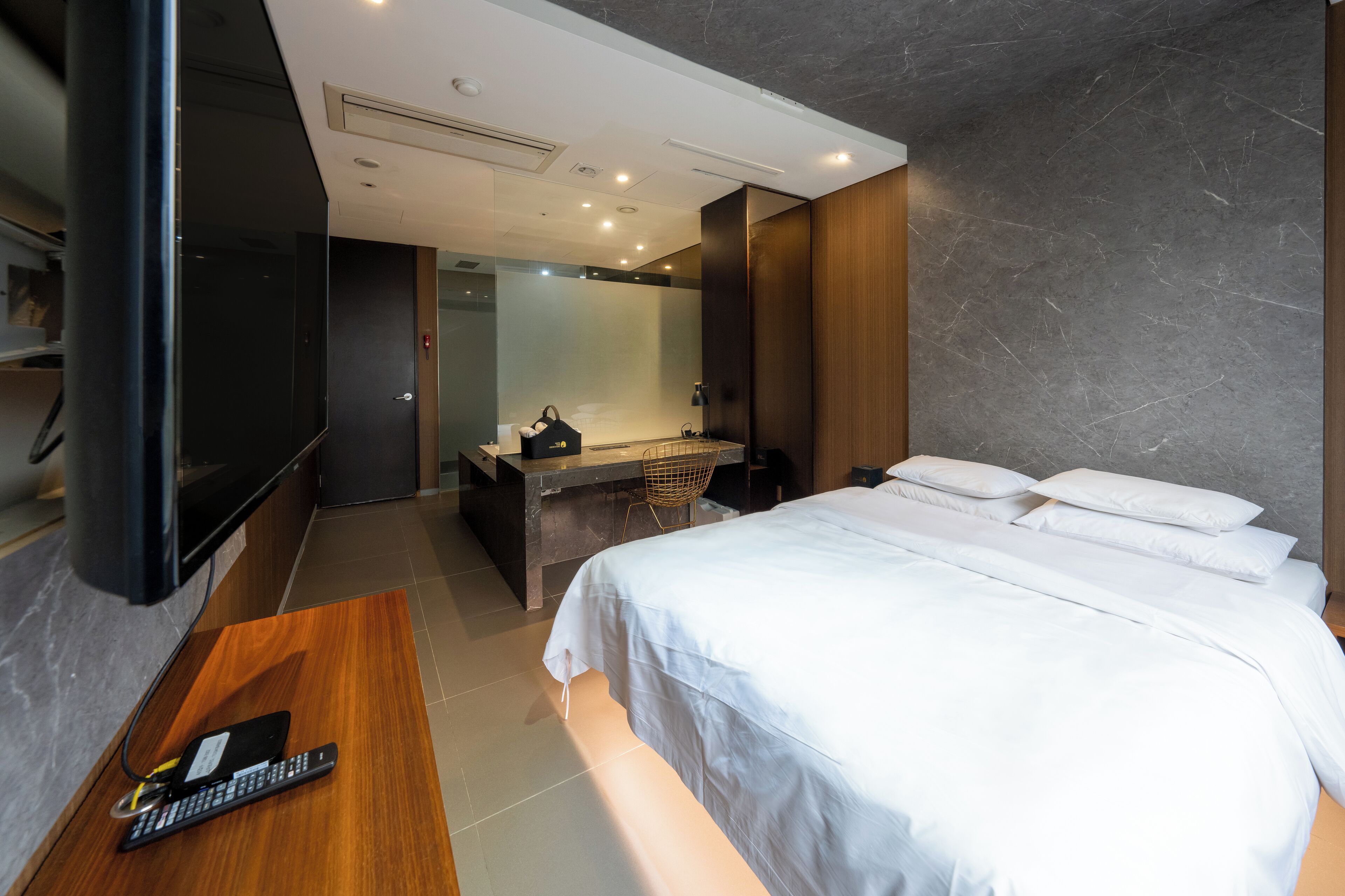 Quarto de hotel moderno com uma cama grande, uma escrivaninha de madeira, uma televisão de tela plana, um banheiro com espelho e uma parede de mármore cinza.