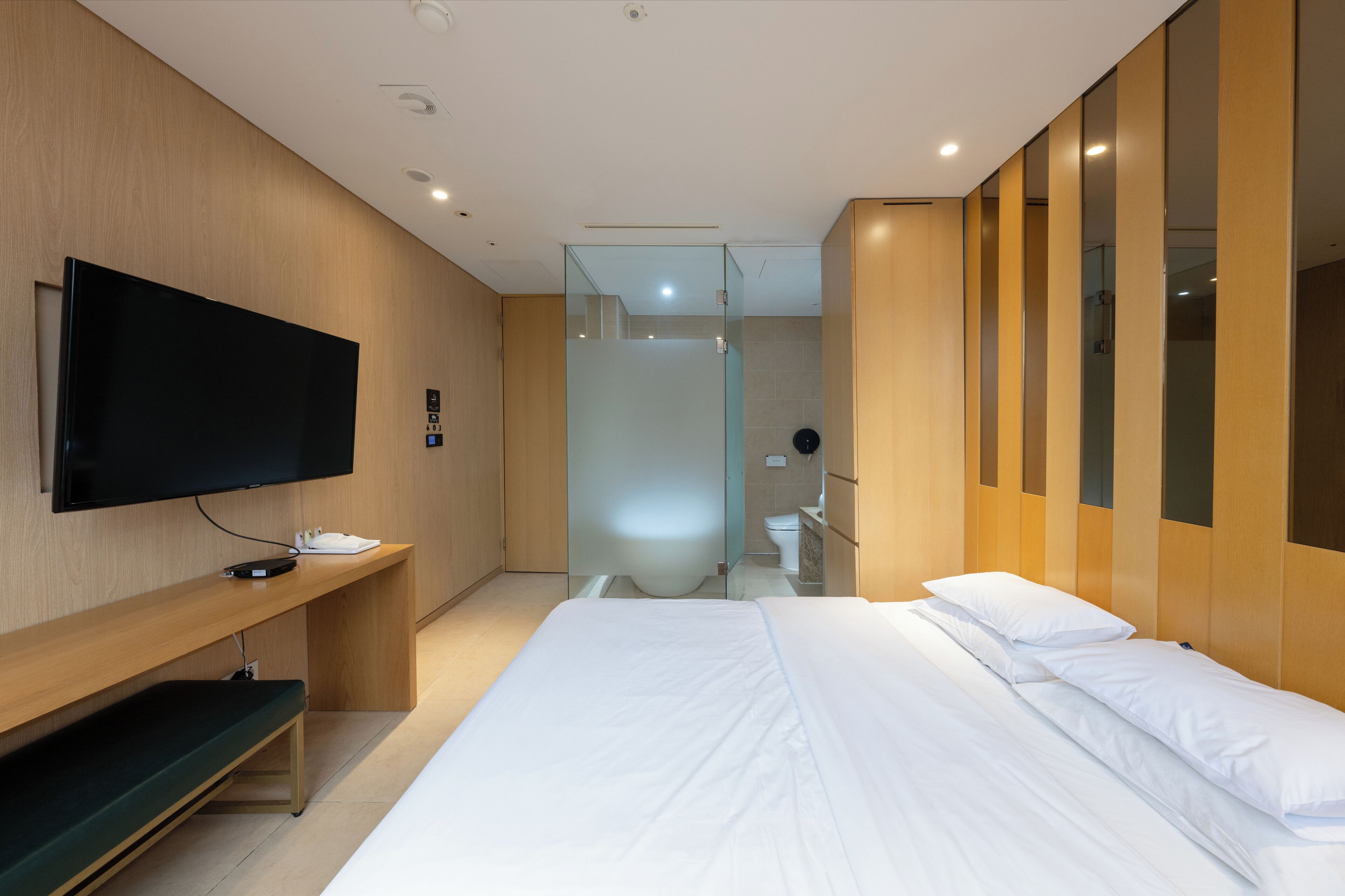 A modern hotel room with a large bed, a flat-screen TV, a wooden bench, and a glass shower enclosure.