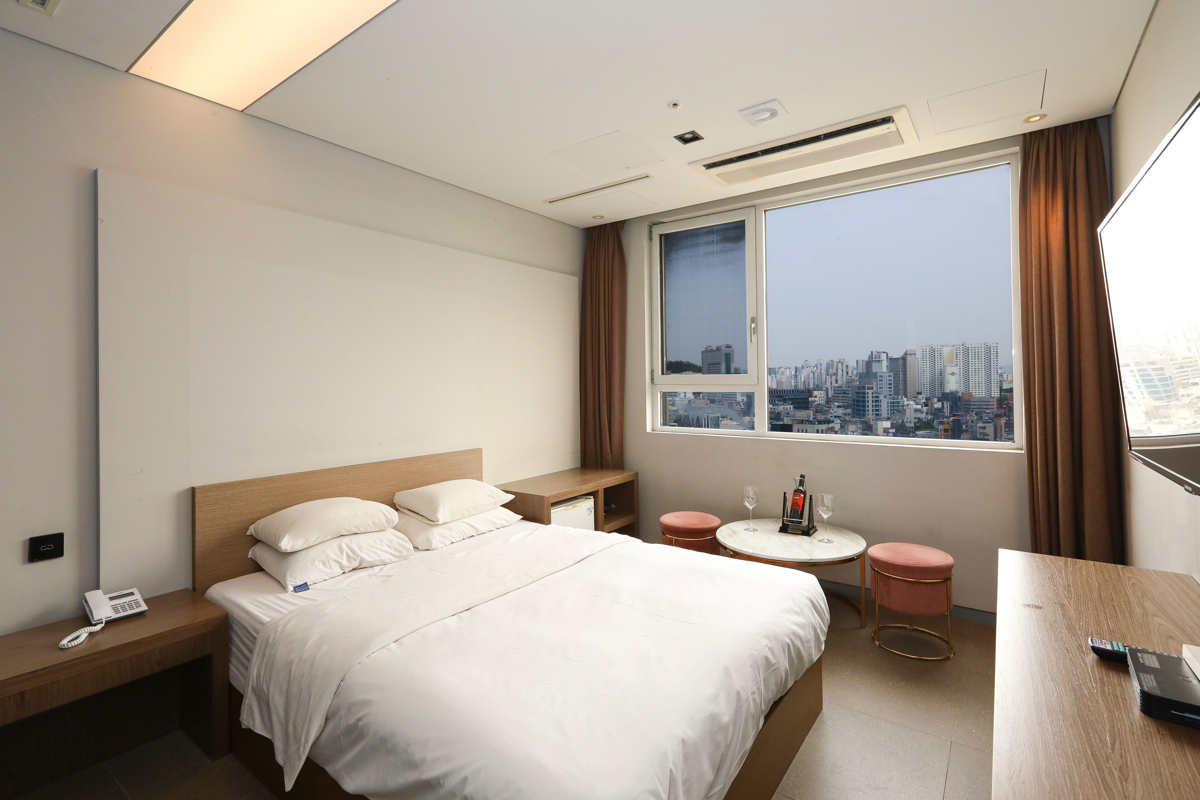 Habitación de hotel con cama, mesita de noche, una pequeña mesa redonda con silla y un ventanal con vista a la ciudad.