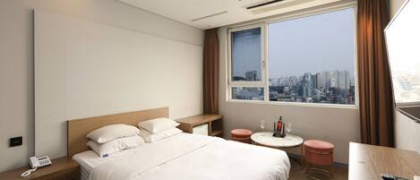 Habitación de hotel con cama, mesita de noche, una pequeña mesa redonda con silla y un ventanal con vista a la ciudad.