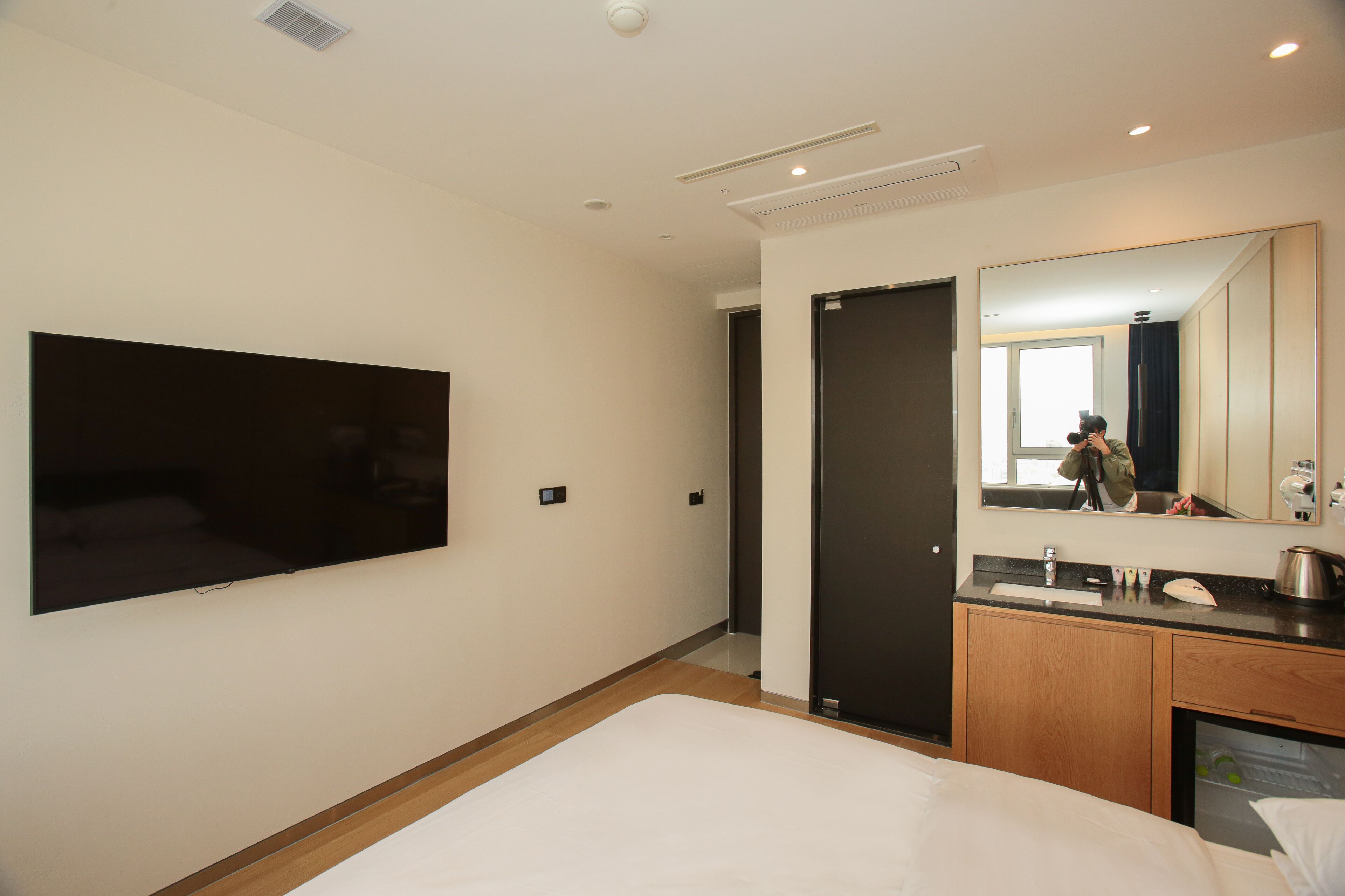 A modern hotel room with a large flat-screen TV mounted on the wall, a wooden floor, a bed with white linens, a wooden cabinet with a sink, and a mirror reflecting part of the room.