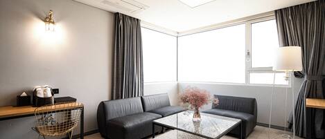 Una habitación de hotel moderna con sofá, una mesa de centro, una lámpara, una pequeña mesa con un florero y una ventana con cortinas.