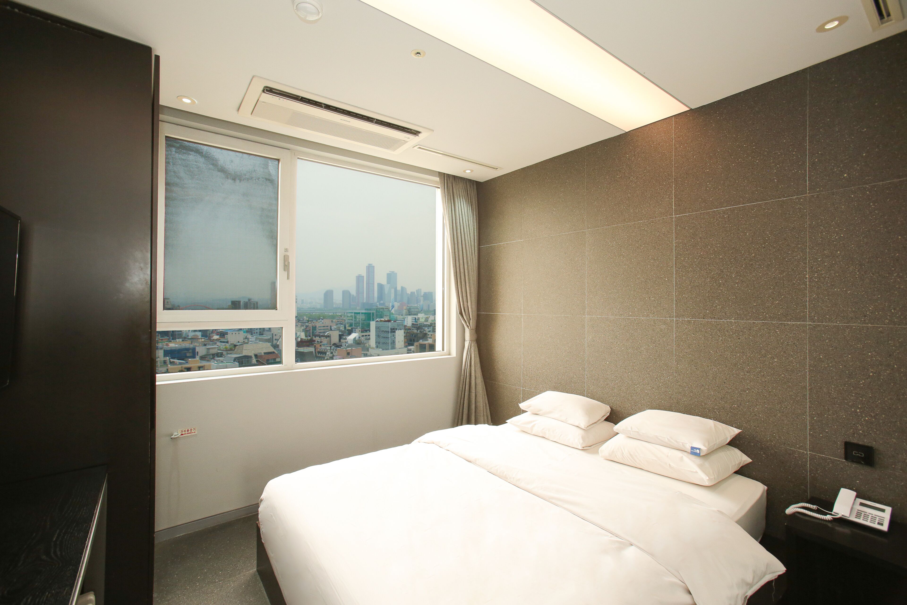 A hotel room with a large bed, a city view, a telephone, and a wall-mounted air conditioner.