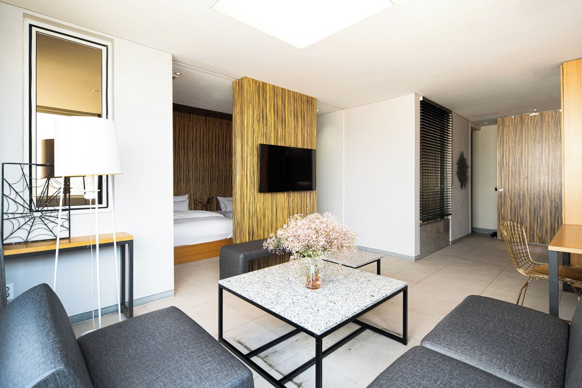 Una habitación de hotel moderna con una cama, un televisor montado en la pared, un comedor con mesa y sillas, y un pequeño rincón de estar con sofás.