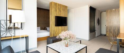 A modern hotel room with a bed, a flat-screen TV, a dining area with a table and chairs, and a decorative vase with flowers.