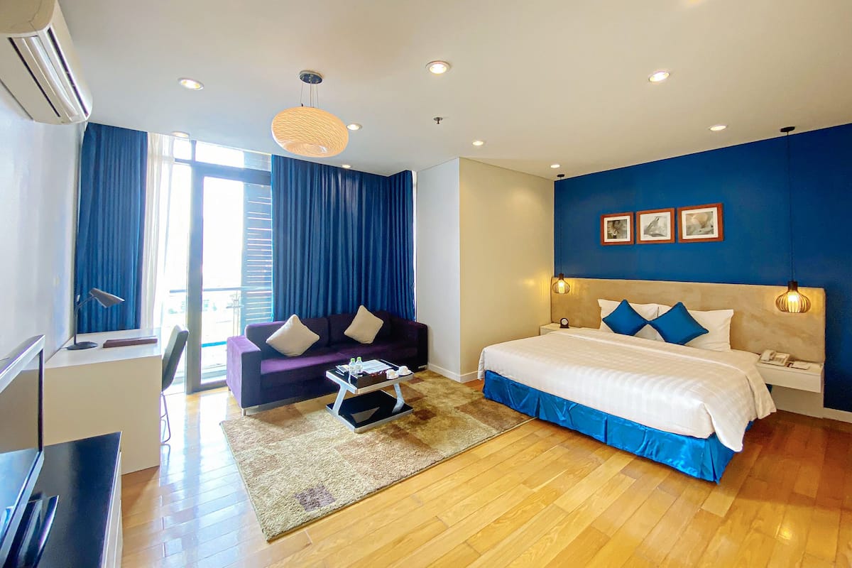 A modern hotel room with a large bed, a sofa, a desk, and a view of the outdoors.